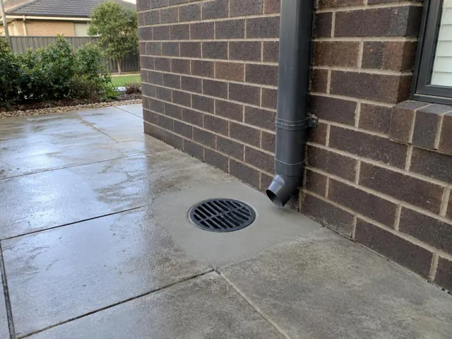 Round outdoor drain grate beside a brick house wall and downpipe on wet paved patio in The Ponds