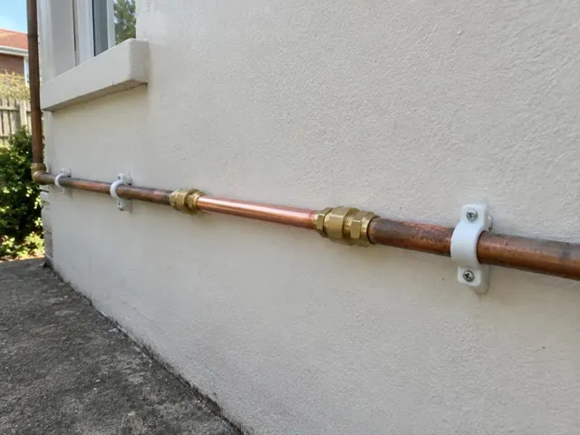 Repaired copper water pipe fixed to an exterior rendered wall beside a window in Riverstone